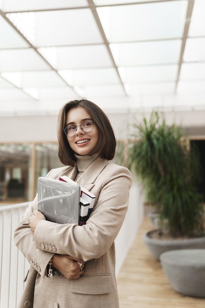 Vertical portrait of young successful woman employee or entrepreneur at office hall tutor carry laptop and notebooks looking away with satisfied pleased smile like her job