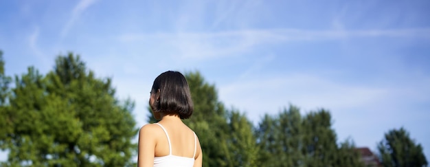 Free photo vertical rear view of young woman doing fitness yoga training in park sitting on rubber mat in