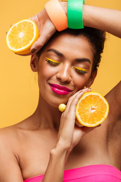 Vertical sensual afro american woman with closed eyes holding two parts of orange and enjoying citrus fruit isolated, over yellow wall