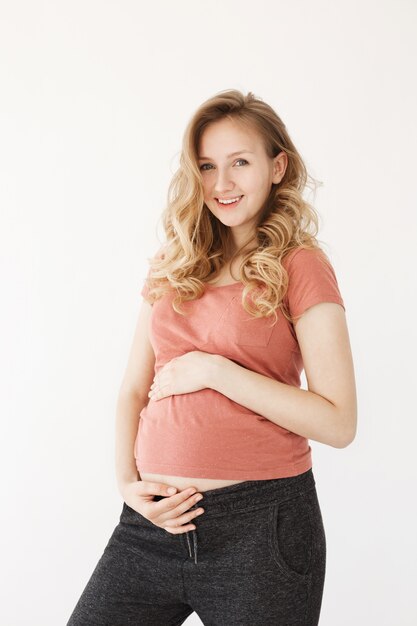 Vertical shor of happy blonde young mother with long hair in sport clothes smiling brightfully, touching pregnant stomach with hands