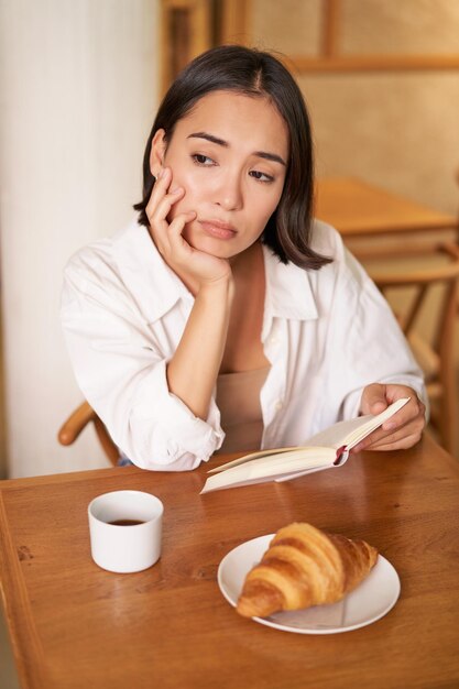 Free photo vertical shot of asian girl sits alone in cafe reads book and looks upset drinks coffee with croissa