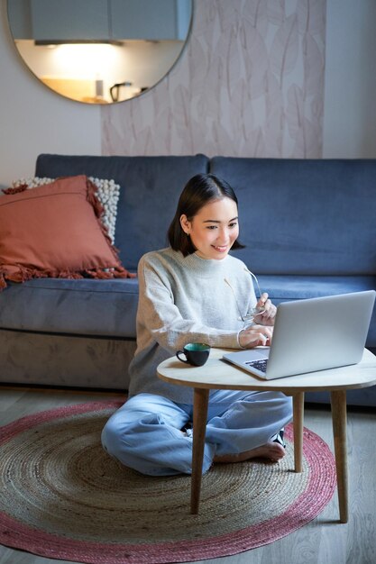 Vertical shot of asian girl sits on floor at home working on laptop studying at cozy place using com