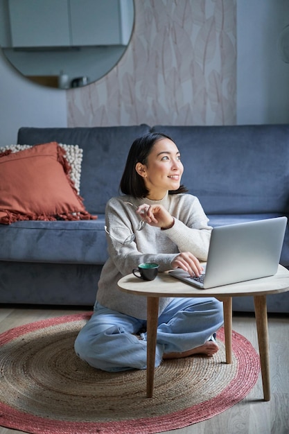 Vertical shot of asian girl sits on floor at home working on laptop studying at cozy place using com