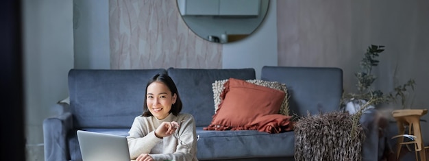 Vertical shot of asian girl sits on floor at home working on laptop studying at cozy place using