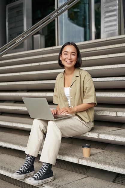 アジアの女の子の垂直ショットは、大学の階段の若い女性学生でラップトップを飲みながらコーヒーを飲みながら座っています