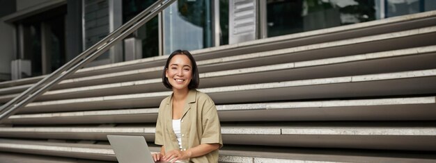 Free photo vertical shot of asian girl sits with laptop drinks coffee on university stairs young woman student