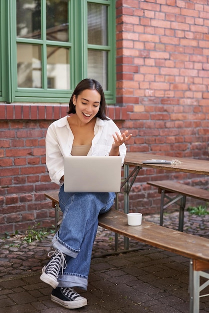 Free photo vertical shot of asian girl with laptop talking on video call chatting online sitting in cafe outdoo