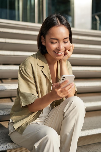 Vertical shot of asian woman student sits on stairs in city looking at mobile phone screen and smiling using smartphone app