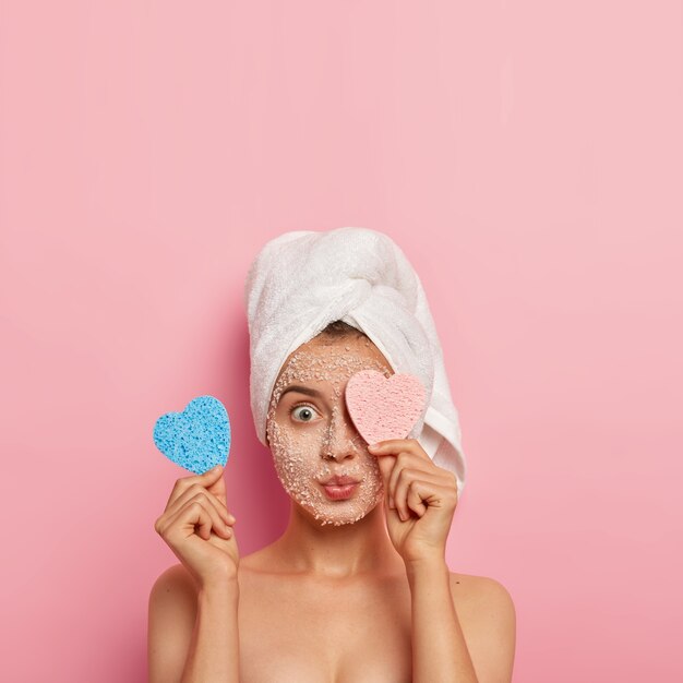 Vertical shot of astonished young woman covers one eye with cosmetic sponge, has morning face treatment, applies white sea salt mask for flawless skin, absorbes nutrients