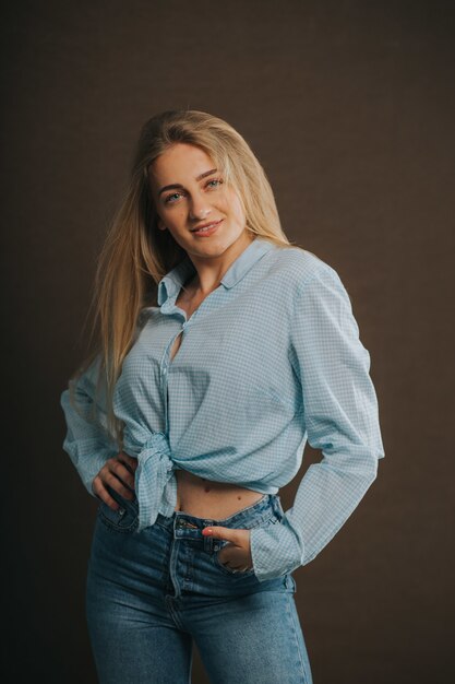 Вертикальный снимок привлекательной блондинки в джинсах и короткой рубашке, позирующей на коричневой стене