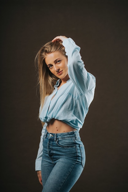 Free photo vertical shot of an attractive blonde female in jeans and a short shirt posing on a brown wall