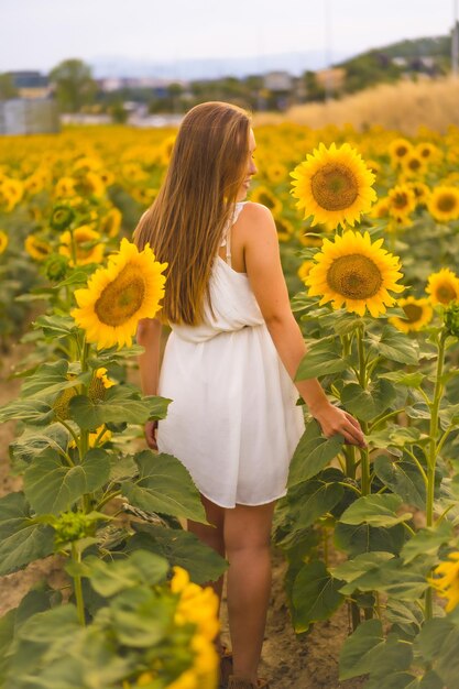 Vertical shot of an attractive blonde female in a white dress posing in a sunflower field