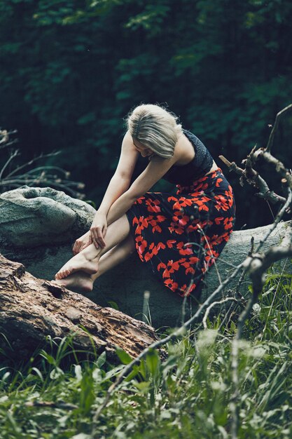 Vertical shot of an attractive blonde female with a floral dress sitting on a tree log looking down