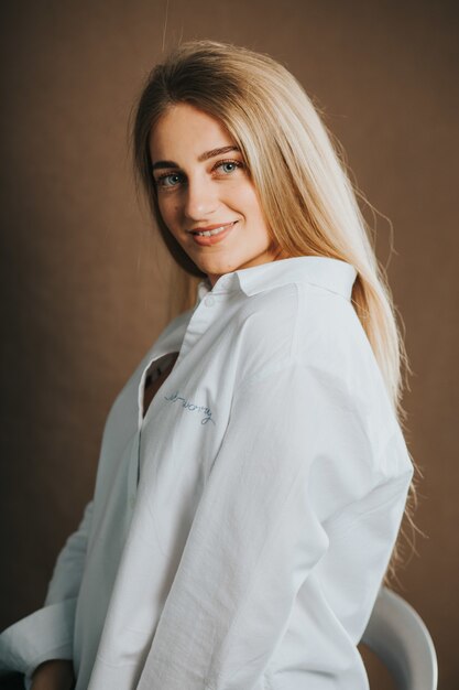 Вертикальный снимок привлекательной блондинки в белой рубашке, позирующей на коричневой стене