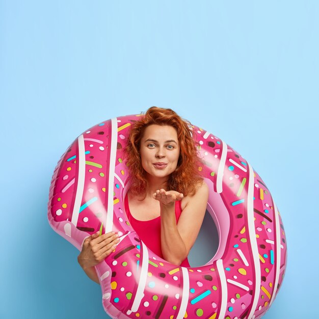 Vertical shot of attractive female model sends air kiss, dressed in swim suit, stands with rubber donut, expresses love, has summer holiday