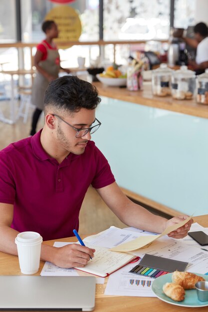 Vertical shot of attractive hipster student prepares financial project, rewrites information from document in notepad, sits at desk in cozy restaurant, wears eyewear, poses indoor. Paperwork concept