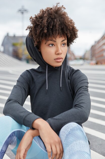 Vertical shot of attractive thoughtful African American hipster wears hoodie, sits on asphalt