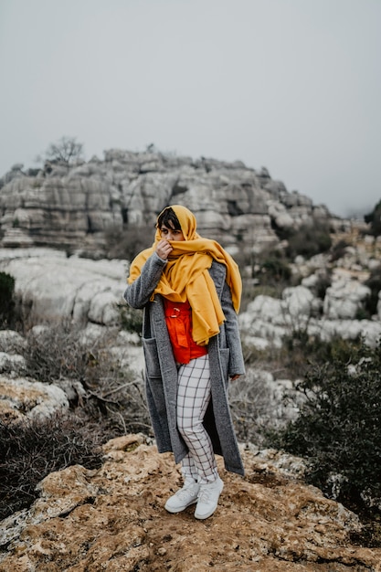 Vertical shot of an attractive young female with a coat and a red scarf posing on top of a cliff