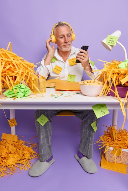 Free photo vertical shot of bearded elderly man works in home office holds mobile phone in hand listens music via headphones dressed in domestic clothes makes list to do on sticky notes sits at table indoor