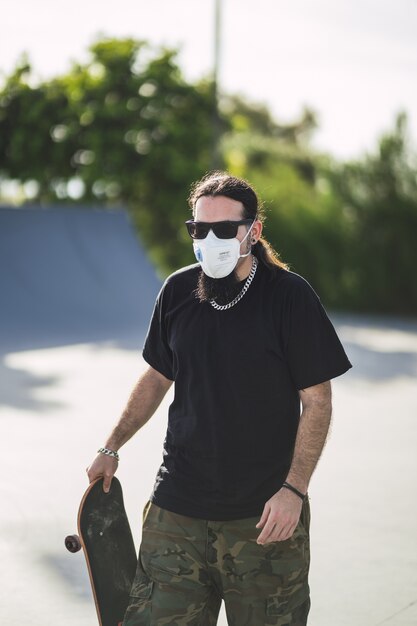 그의 스케이트 보드를 들고 공원에서 산책하는 얼굴 마스크를 쓰고 수염 난 남성의 세로 샷