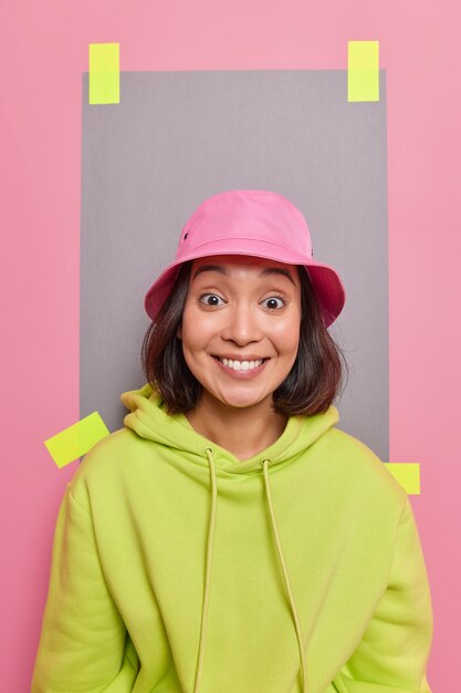 Вертикальный снимок красивой азиатской женщины с темными волосами, зубастой улыбкой, в розовой панаме и зеленой толстовке, радостно выглядящей из-за оштукатуренного листа бумаги в помещении.