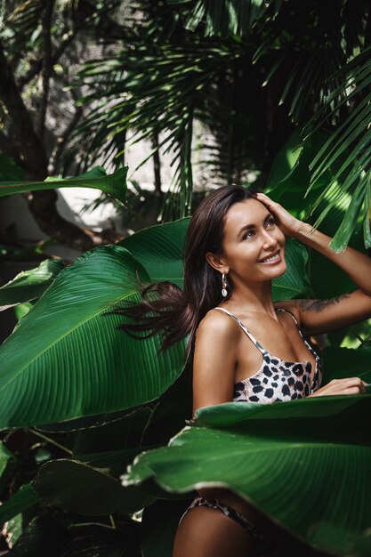 Vertical shot of beautiful brunette female posing near palm leaves in a bikini, hair flying in the air.