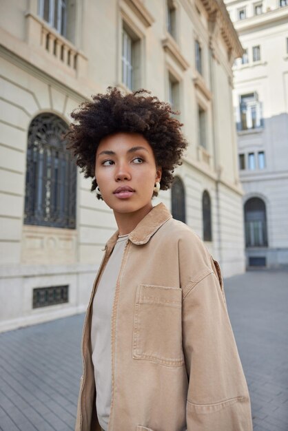 Vertical shot of beautiful brunette woman wears casual beige jacket concentrated into distance walks around ancient city buildings thinks about something strolls outdoors explores new places