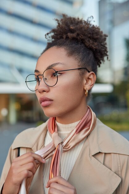 Vertical shot of beautiful curly haired millennial gir wears round spectacles for vision correction coat and tied kerchief around neck looks thoughtfully forward while strolling outside during daytime
