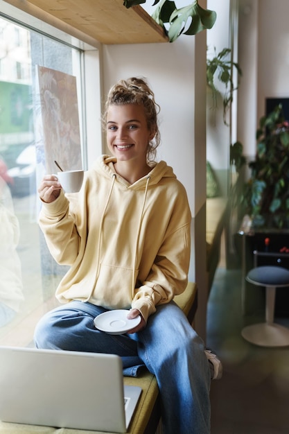 Вертикальный снимок красивой девушки, сидящей у окна кафе, пьющей кофе и смотрящей на улицу, счастливо работающей на ноутбуке Студентка наслаждается напитком и учится удаленно от кофейни