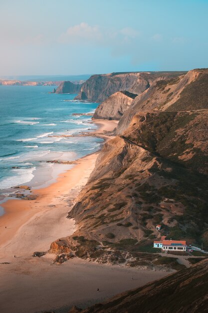 Vertical shot of the beautiful ocean by the rocky cliff and mountains under the clear blue sky