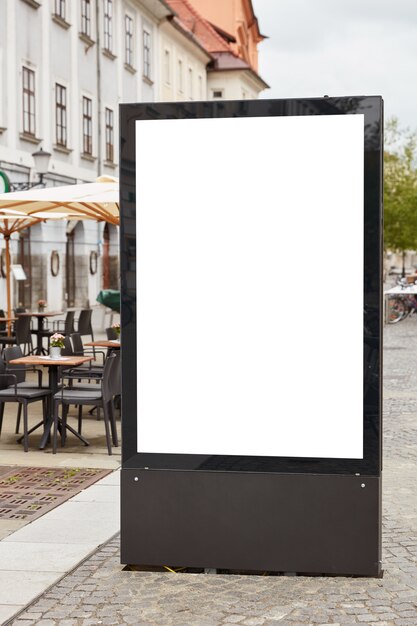 Vertical shot of blank billboard stands on pavemenet against city background near outdoor cafeteria