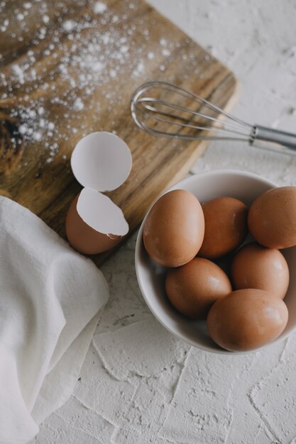 Vertical shot of a bowl of eggs and a silver whisk next to a chopping board on a white surface