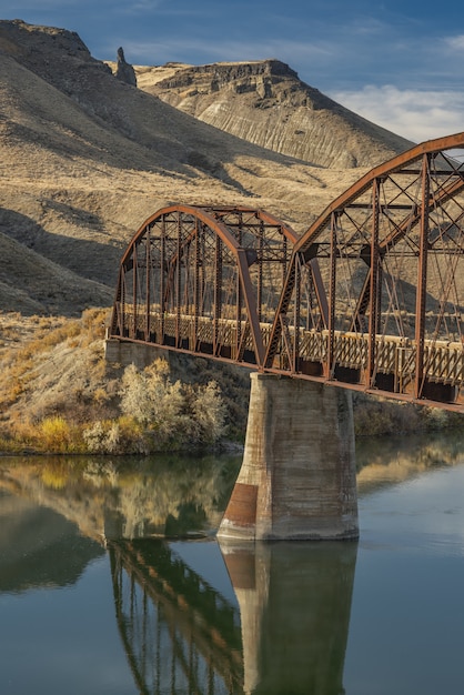 Vertical shot of a bridge over the river with mountains and a blue sky