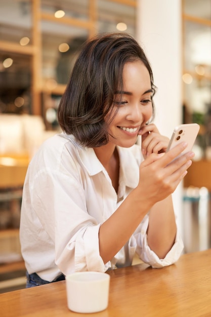 Vertical shot of brunette asian girl sitting in cafe using smartphone app chatting on mobile phone a