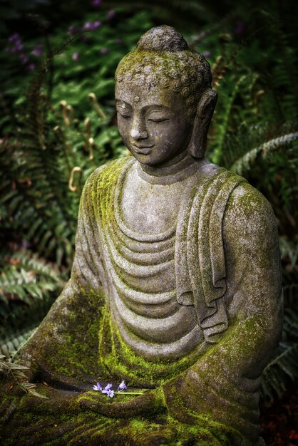 Vertical shot of a Buddha statue with moss on top and greenery on the distance