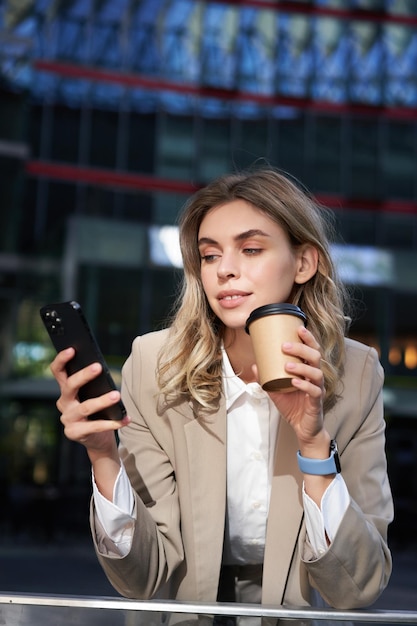 Vertical shot of businesswoman drinks coffee looks at mobile phone app corporate woman on her lunch