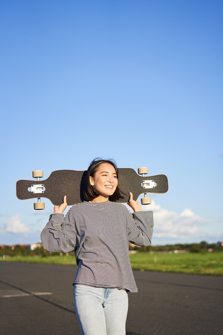 免費 Photo 一位无忧无虑的亚洲女孩与长板年轻女滑板运动员的垂直镜头,她的肩膀上拿着巡洋舰