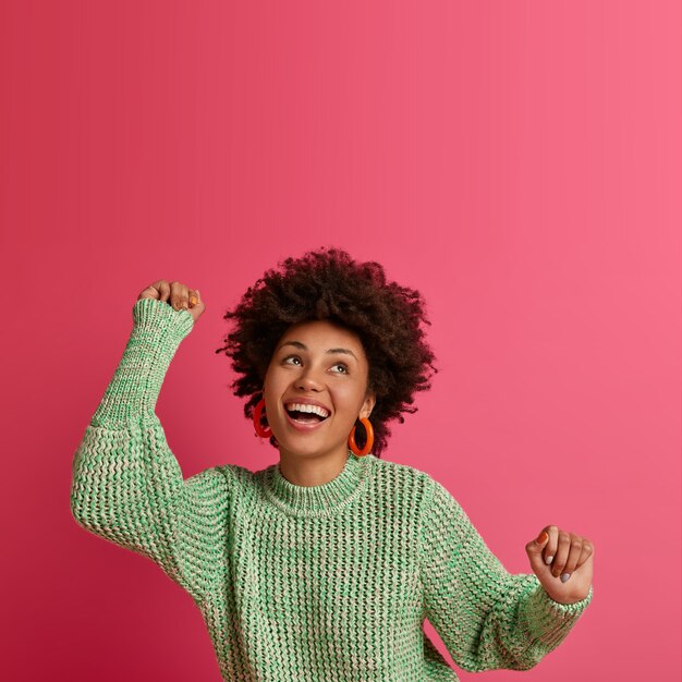 Free photo vertical shot of carefree young woman does champion dance, looks with broad smile, concentrated above, enjoys perfect day off, feels like winner, wears warm jumper, isolated on pink wall