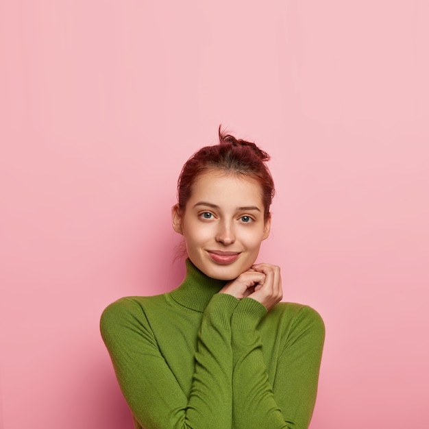 Colpo verticale di affascinante donna europea tiene le mani vicino al viso, indossa dolcevita verde casual, guarda direttamente la telecamera, isolato su sfondo rosa, spazio libero per il tuo