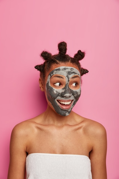 Free photo vertical shot of cheerful african american woman has fresh clean skin, looks aside, applies clay mask, enjoys facial treatment, stands wrapped in bath towel, isolated on pink wall. beauty face care