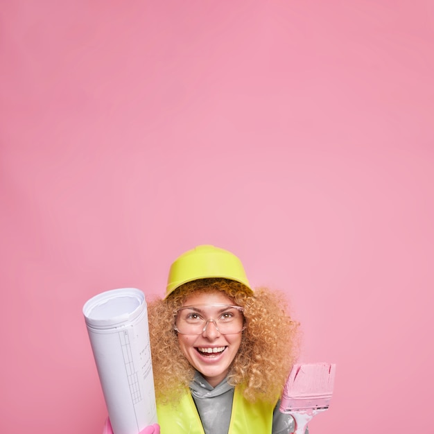 Vertical shot of cheerful curly woman wears hardhat transparent glasses holds blueprint and paint brush comes on construction site 