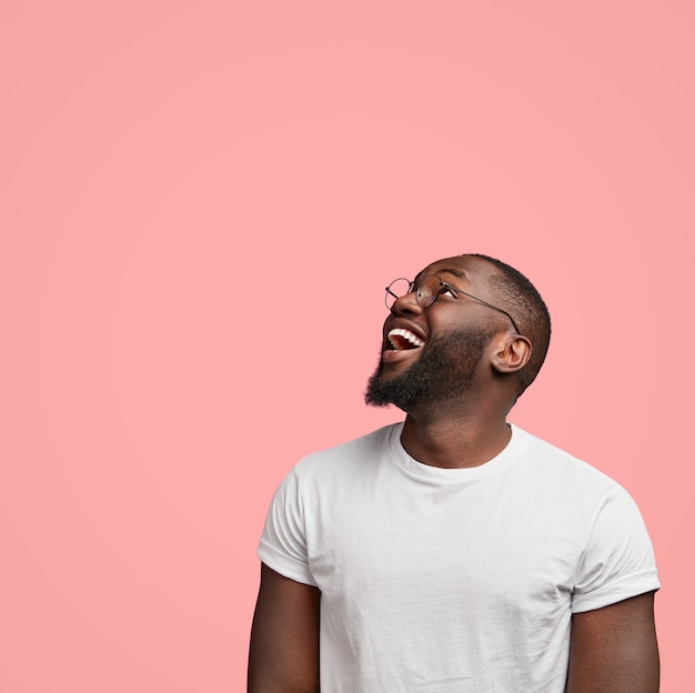 Free photo vertical shot of cheerful dark skinned unshaven man looks positively upwards, being in good mood,