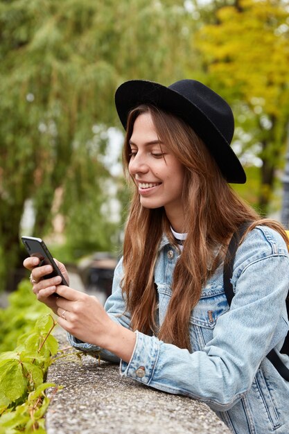 Vertical shot of cheerful fashionable woman reads funny message, uses phone application, poses over green trees outdoor