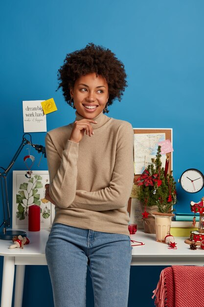 Vertical shot of cheerful satisfied Afro American female student stands near workplace against blue wall., keeps hand under chin and looks away, thinks about future plans pleased work from home