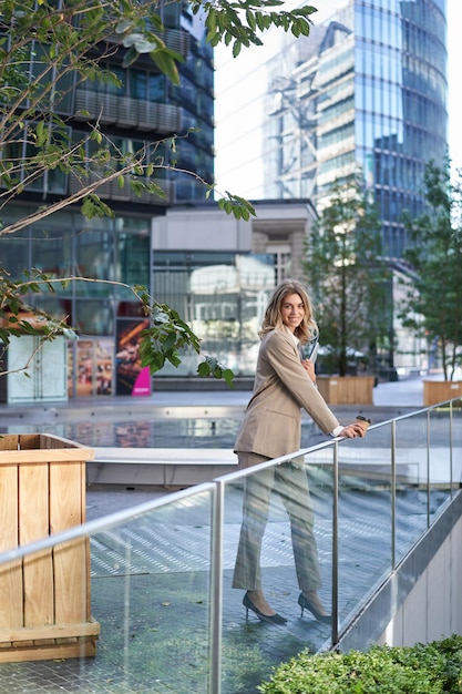 Вертикальный снимок корпоративной женщины в бежевом костюме, стоящей на улице и пьющей утренний кофе