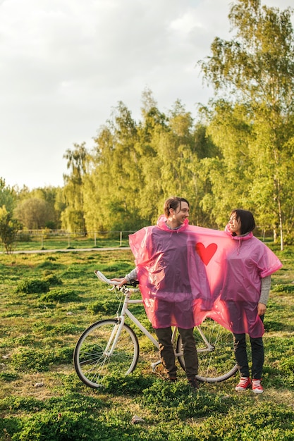 Vertical shot of a couple sharing a pink plastic raincoat at a date with a bike