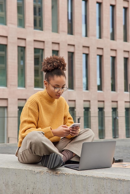 Vertical shot of curly haired millennial girl sits crossed legs uses mobile phone and laptop computer connected to wireless