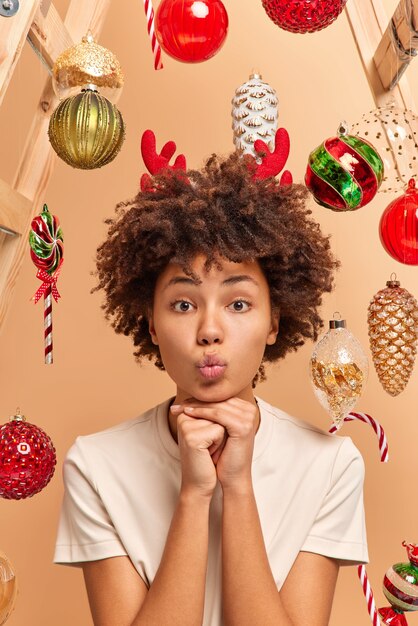 Vertical shot of curly haired woman keeps lips folded and hands under chin looks with romantic expression at camera dressed casually surrounded by christmas toys has festive mood. Holiday celebration