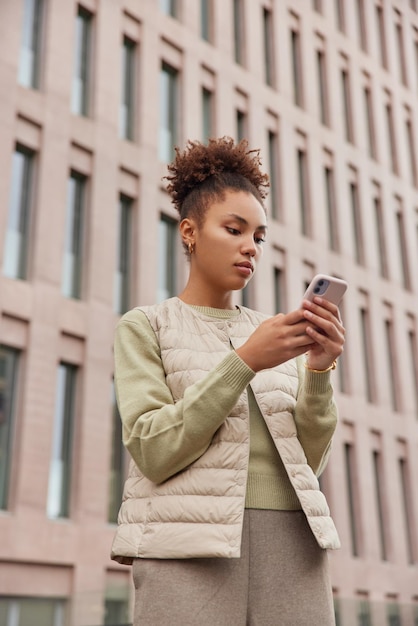 縮れ毛の女性の垂直ショットは携帯電話を使用してウェブサイトを閲覧し、ソーシャルネットワークで自由な時間を過ごしますカジュアルな服を着て現代の都市建設に対抗しますテクノロジーとライフスタイル