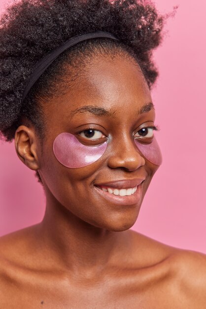 Vertical shot of dark skinned curly female model applies hydrogel patches under eyes for rejuvenation